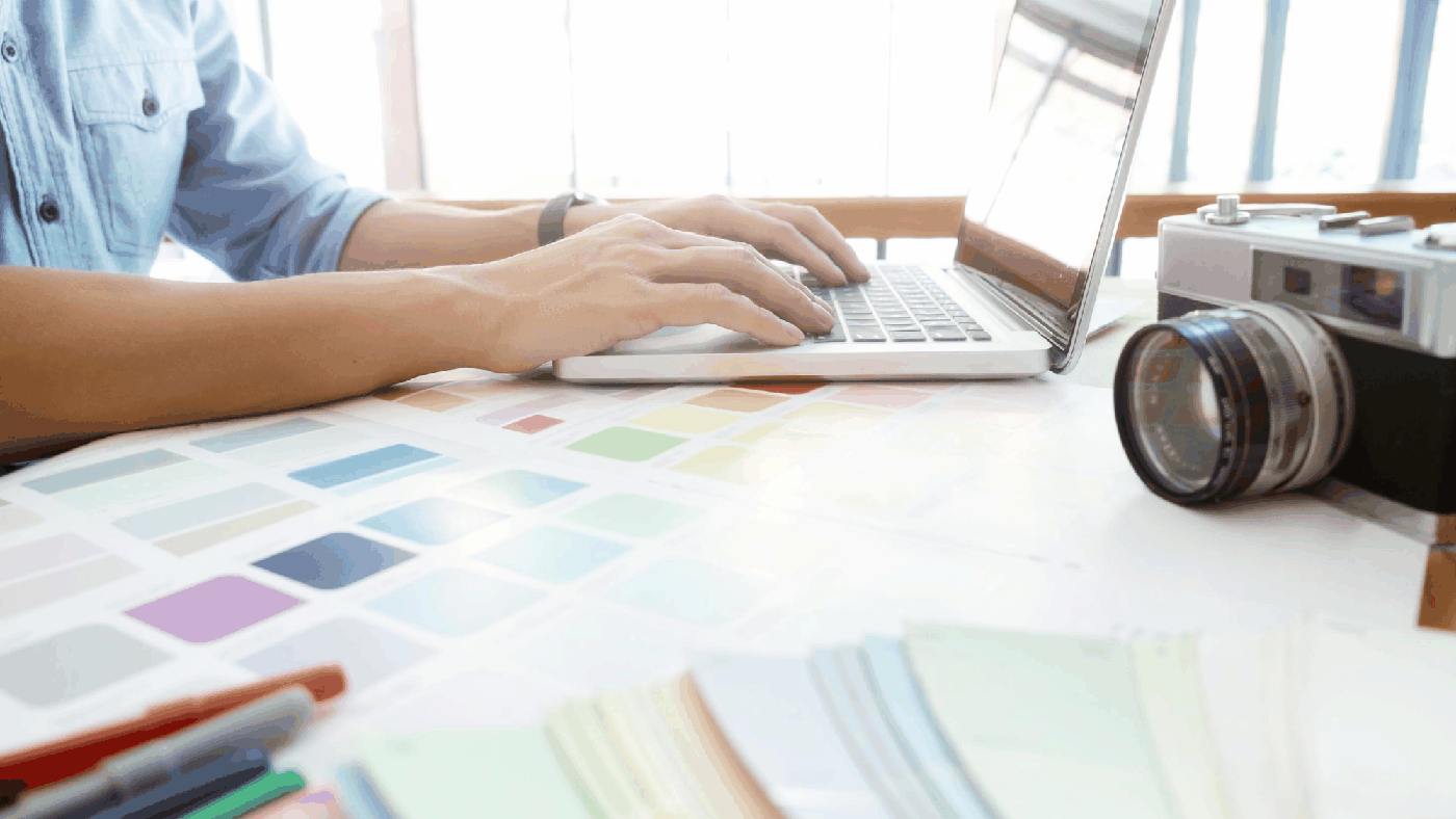 Hands typing on a laptop beside printed colour swatches and a film camera